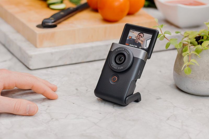 A Canon PowerShot V10 stands on a kitchen worktop with a chopping board in the background and a vlogger's hand resting on the surface in front of it.
