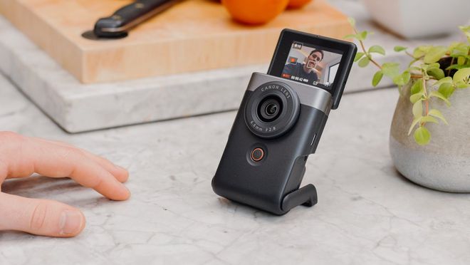 Et Canon PowerShot V10 står på en køkkenbordplade med et skærebræt i baggrunden og en vloggers hånd, der hviler på overfladen foran den.