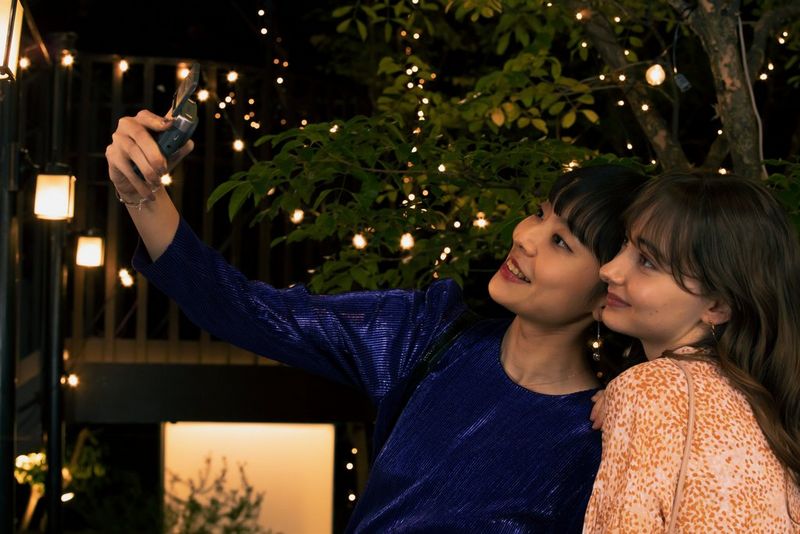 Two women film themselves with a Canon PowerShot V10 vlogging camera in front of a Christmas tree adorned with fairy lights.