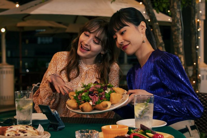 Two women seated at a restaurant table, holding up a plate in front of a Canon PowerShot V10 on its built-in stand.