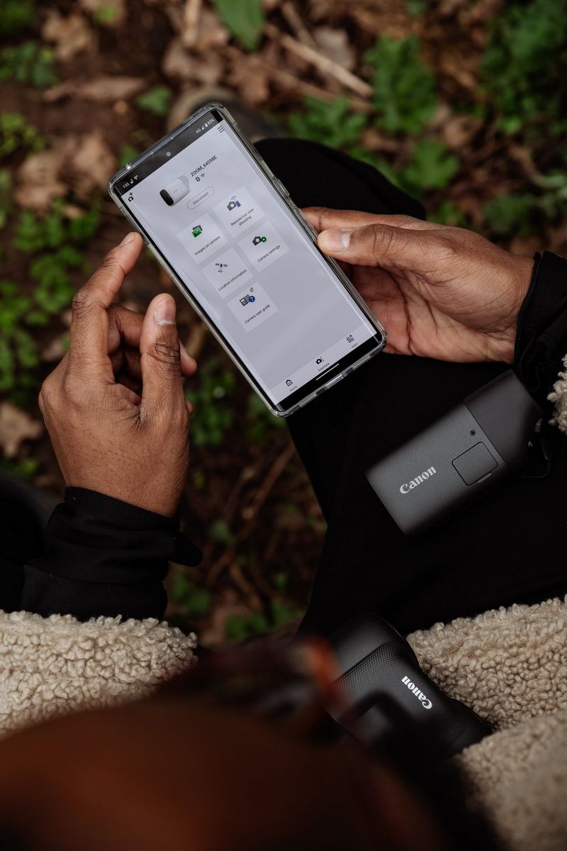 A pair of hands holds a smartphone next to a Canon PowerShot ZOOM, showing it connected using the Canon Camera Connect app.