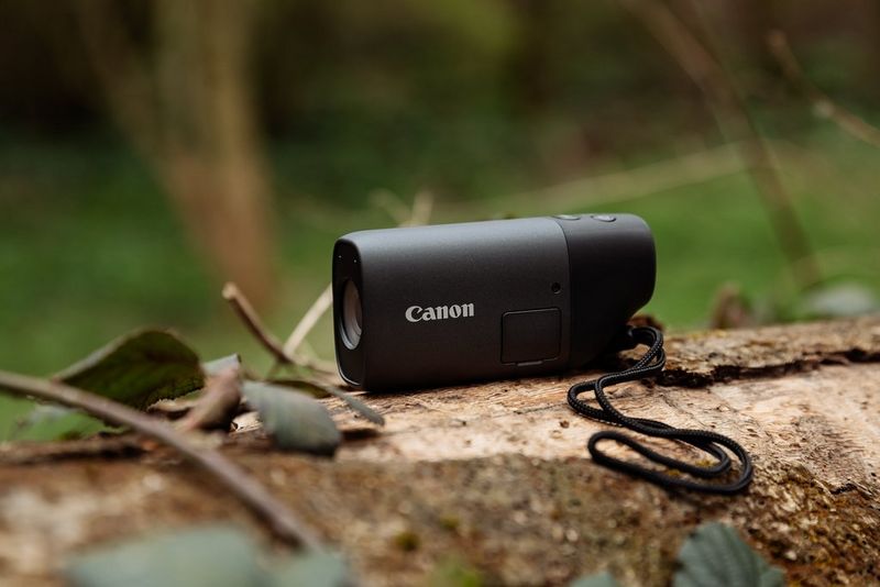 A Canon PowerShot ZOOM camera sits atop a fallen tree trunk, wooded landscape out of focus in the background.