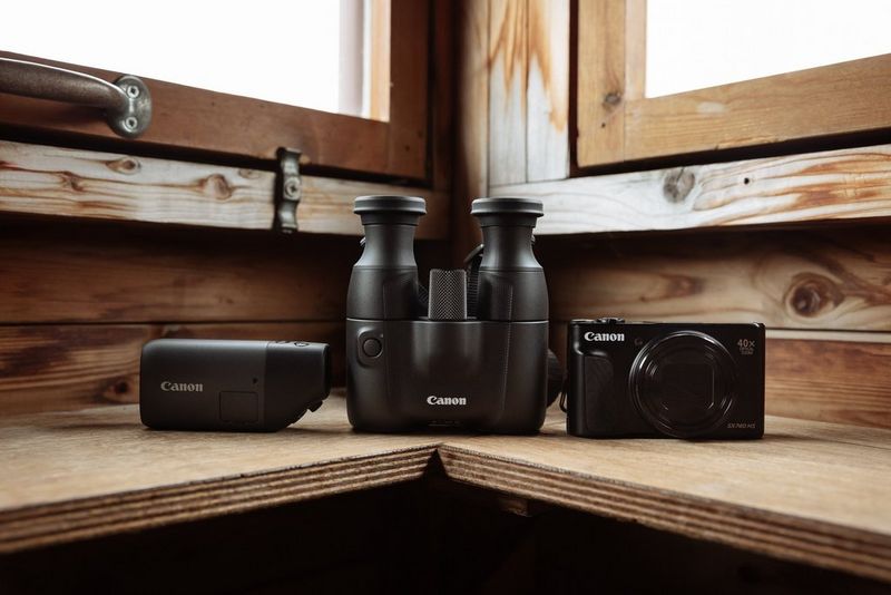 A shot of a Canon PowerShot ZOOM, Canon binoculars and a Canon camera resting on a wooden bench in a wildlife hide.