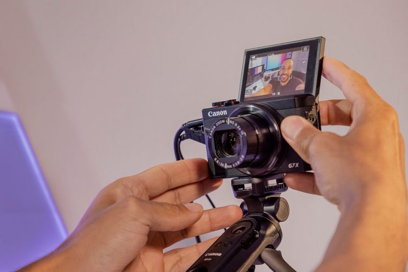 Hands adjust the LCD screen on a Canon PowerShot G7 X Mark III on a tripod, facing it forward so the person can see themselves as they're filming.