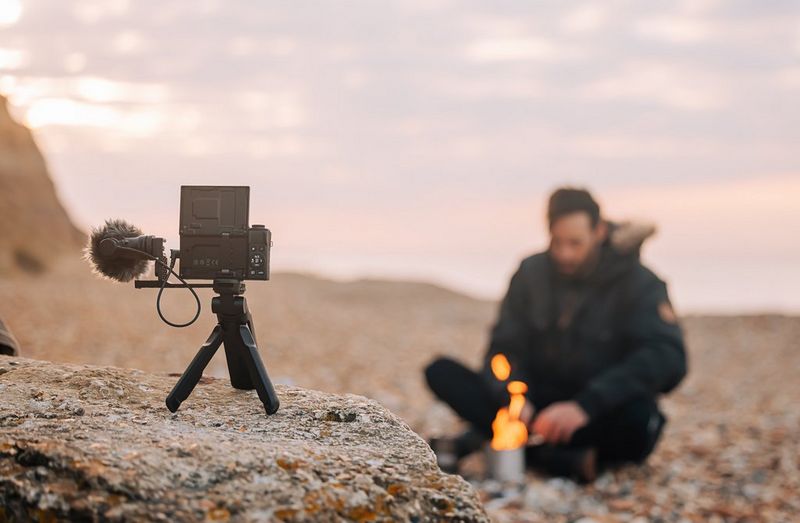 A man sits on a pebbled beach in front of a small fire. A Canon PowerShot G7 X Mark III is set up on a tripod with a microphone on a rock in front of him.