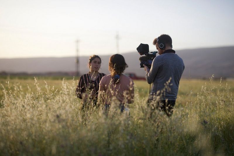 A man holding a Canon EOS C70 camera films a woman standing in a field of long grass. Another woman stands beside him.