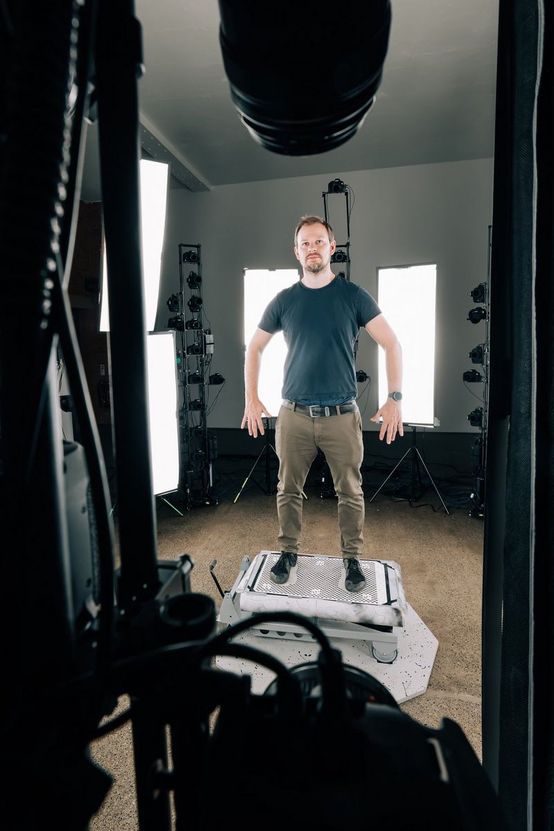 A man in t-shirt and trousers stands on a scissor lift in the middle of a circle of lights and camera arrays.