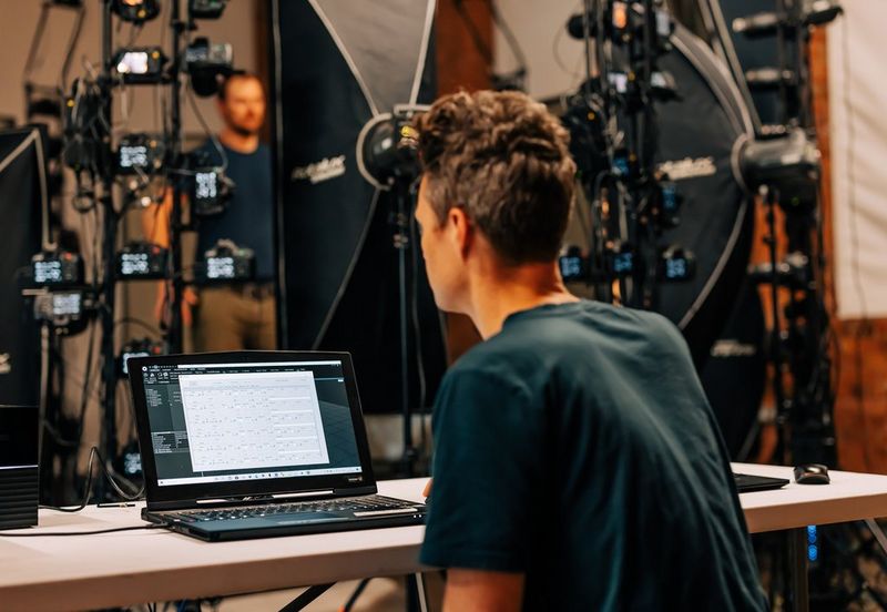 A technician sits at a laptop while a man stands within the 3D photogrammetry array in the background. 