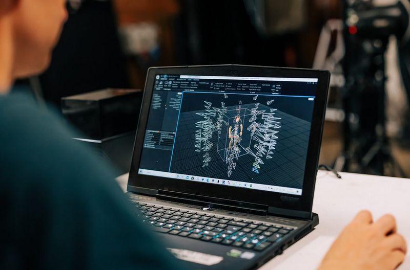 A laptop with a 3D model on its screen of a standing figure surrounded by representations of cameras. 