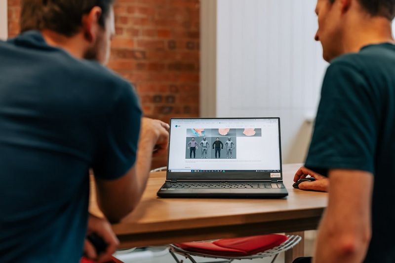 Two men sit at a table looking at a laptop screen showing 3D models of human figures.