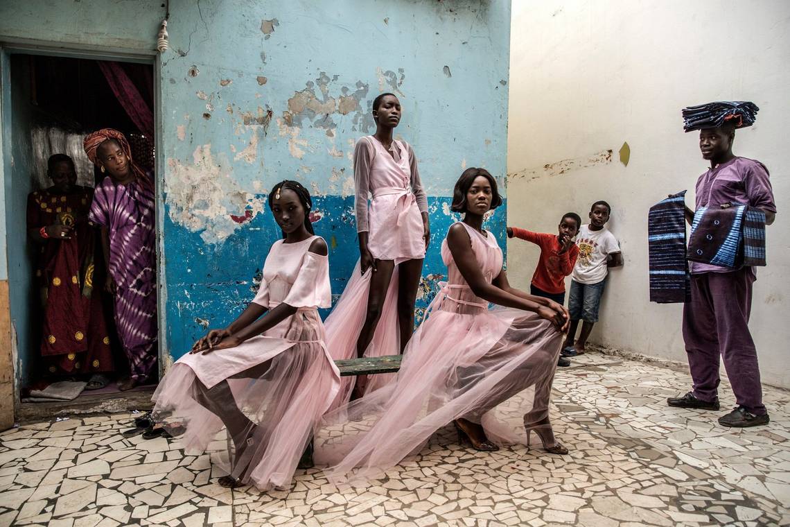 Three Senegalese models in flowing pink dresses show off a fashion line in front of a building in Dakar with flaking blue walls.