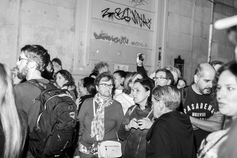 A large group of people stand near a graffitied concrete wall, shot in black and white. 