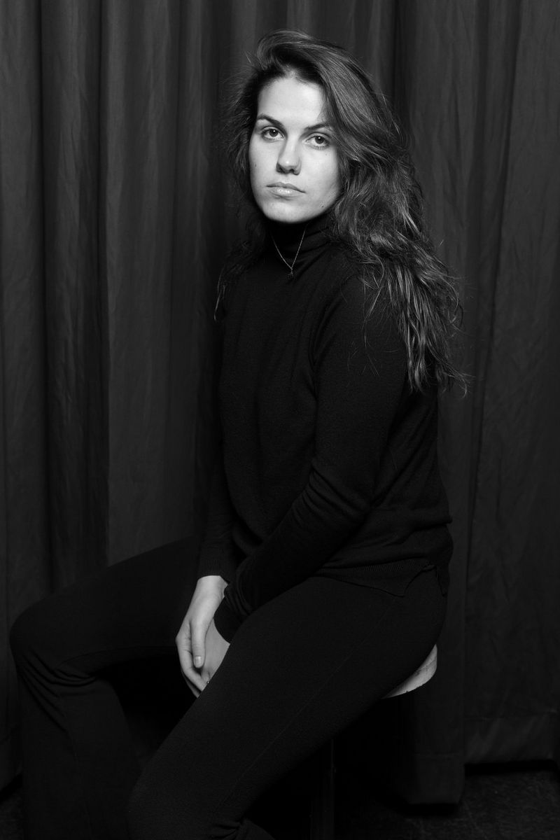 A black and white photo of photographer Bebe Blanco Agterberg, dressed in black, sitting on a stool and staring at the camera.