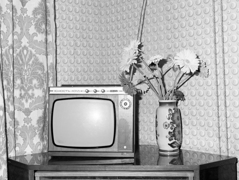 An old-fashioned TV and vase of flowers on a corner table, against patterned wallpaper and curtains. Photographed in black and white.
