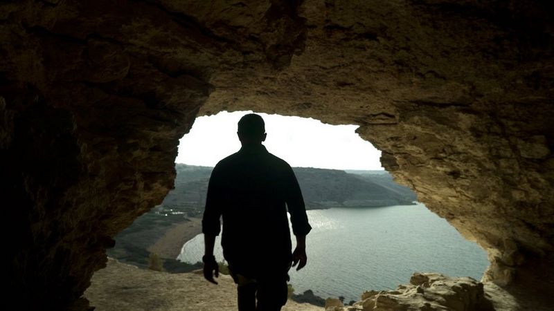 A figure is silhouetted against the entrance to a seaside cave in a still image from Martin Christ's award-winning docudrama The Mystery of the Trojan Horse.