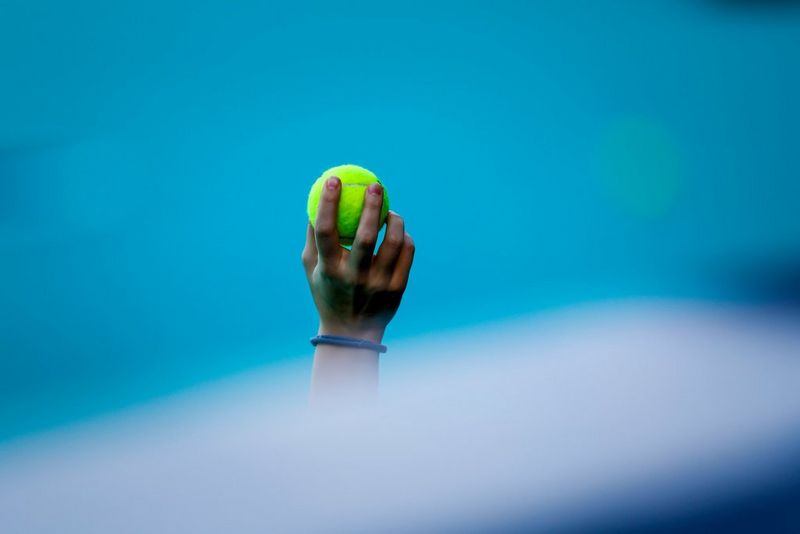 A hand holding a tennis ball is captured in focus against an out-of-focus blue background. Taken on a Canon EOS R3 with a Canon RF 400mm F2.8L IS USM lens.