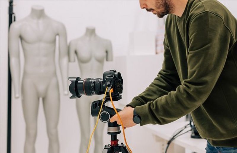 A man in a fashion studio adjusts a Canon EOS R5 camera on a tripod, with two mannequins in the background.