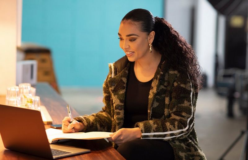 Director Ashleigh Jadee writes in her notebook while seated at a desk in front of an open laptop. She is wearing a green camouflage fleece jacket and smiling at the screen.