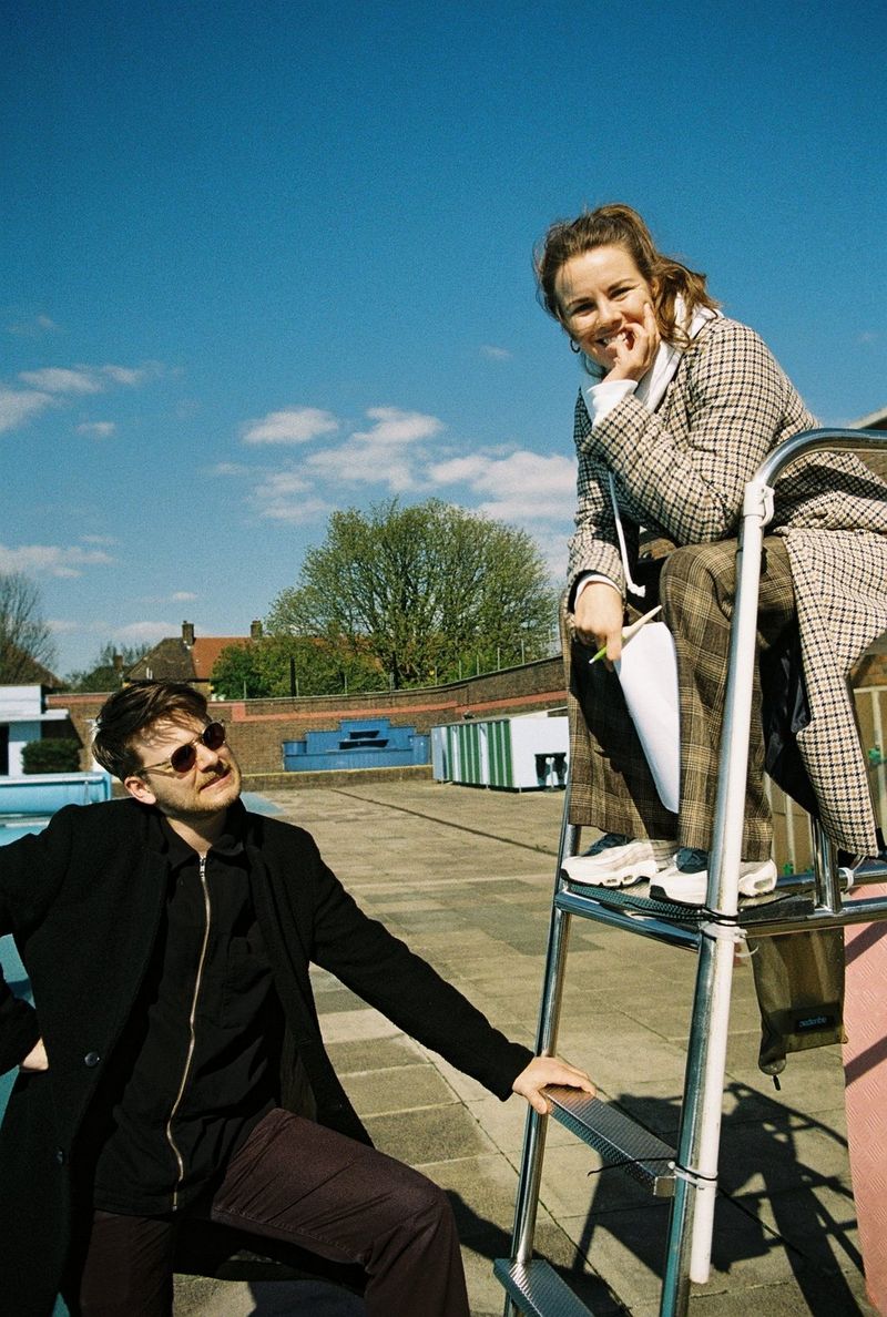On a sunny day, filmmaker Basha de Bruijn sits on the top step of a stepladder, while a man in a long black coat and sunglasses looks up at her.