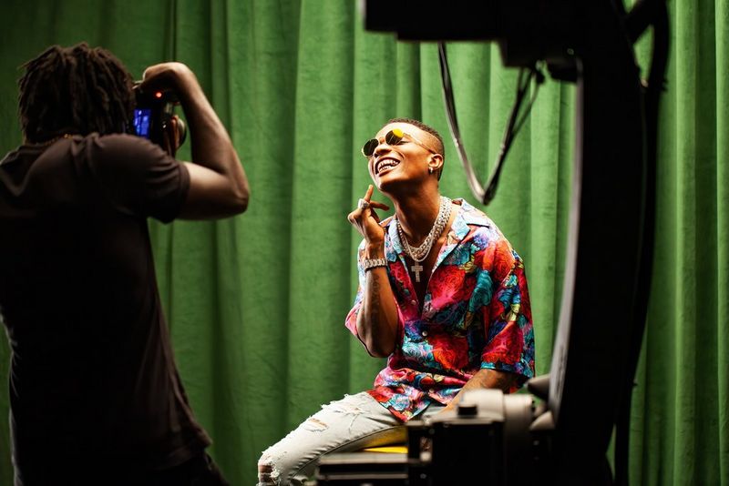 Nigerian singer Wizkid, wearing a colourful shirt and sunglasses, smiles as he has his picture taken against a green curtain.