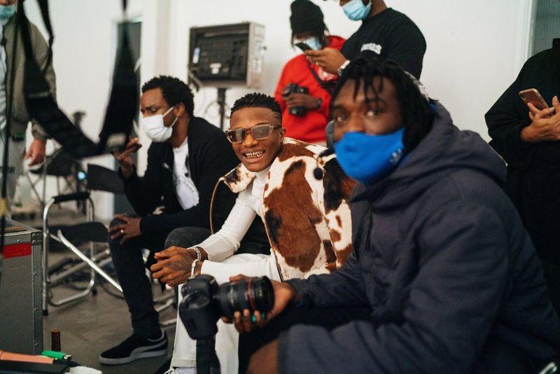 Director Meji Alabi and singer Wizkid take a break on set. Meji is holding a Canon camera and wearing a blue mask. They are surrounded by crew members.