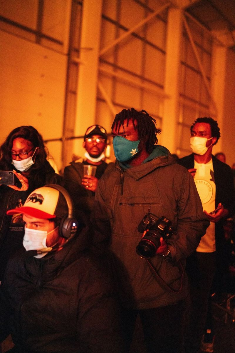 A masked Meji Alabi holds a Canon camera on an outdoor set surrounded by his crew.