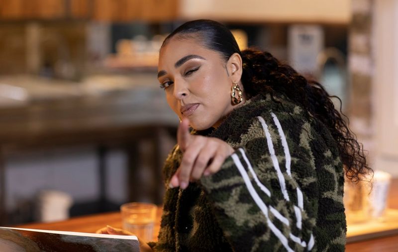 Director Ashleigh Jadee points and winks at the camera. She is seated at a table and wearing a green camouflage fleece jacket and gold hoop earrings.