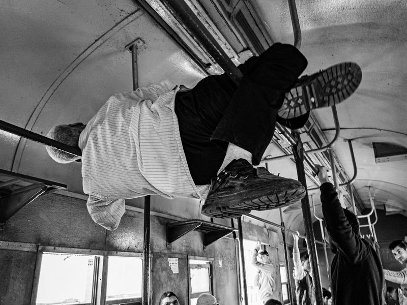 A black and white photograph of a man, seen from behind, balanced across the top handrails of a rickety train carriage. Other passengers can be seen holding on and looking at the man. Taken by Ahmed Qabel.