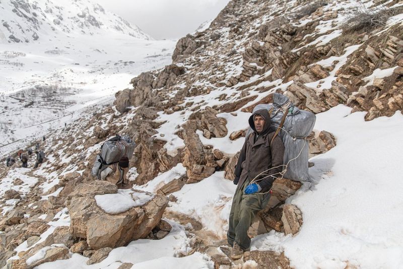 A man with a large, full sack strapped to his back rests against rocks on a snowy mountainside, in a photograph by Ebrahim Alipoor.