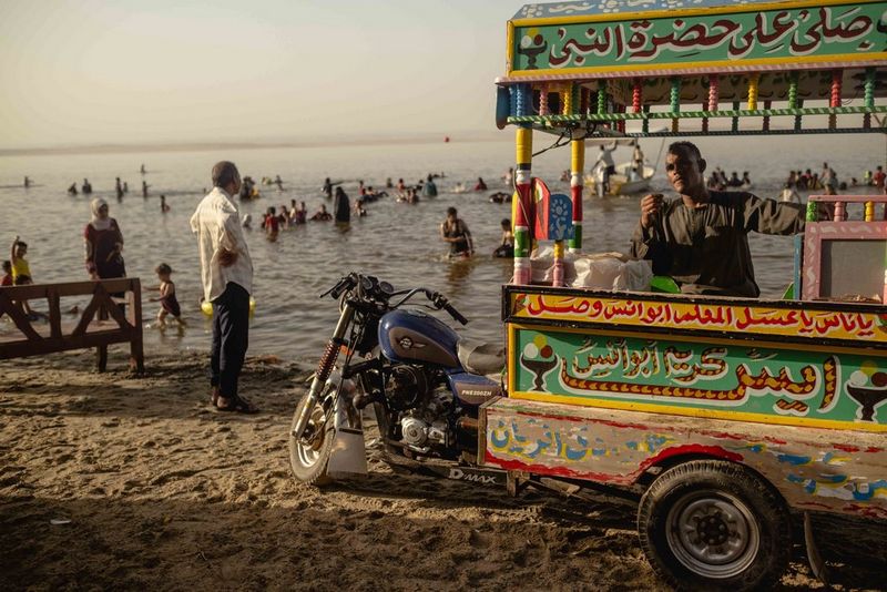 An ice cream cart attached to a motorcycle is parked on the sandy beach at the edge of a lake. Holidaymakers can be seen behind playing and splashing in the dirty-looking water. Photograph by Fatma Fahmy.