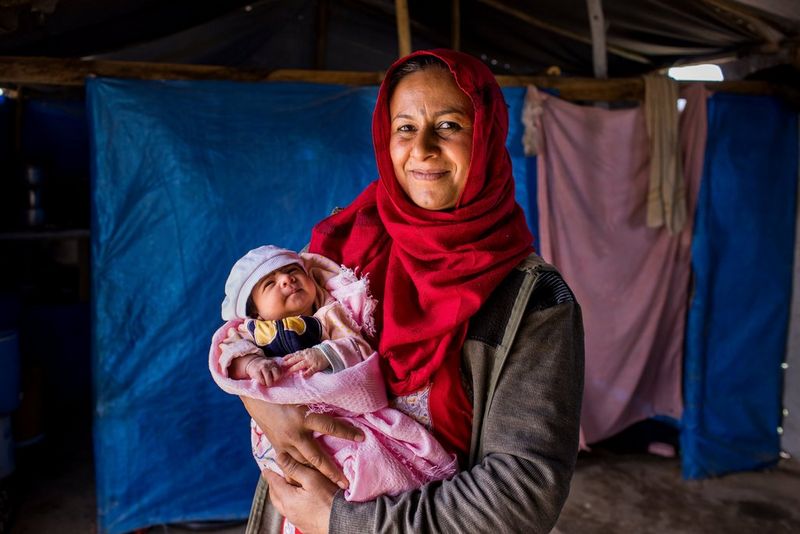 A woman in a red headscarf stands smiling at the camera, holding a baby wrapped in a pink blanket in her arms. 