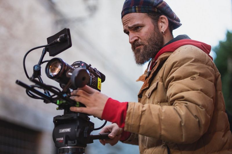 Cinematographer Ivan D'Antonio, wearing a light brown jacket and tartan cap, using a Canon Cinema EOS camera outdoors.