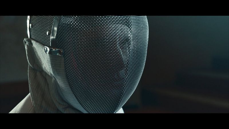 A close-up of a male fencer wearing an epee mask staring intently. The image has been colour graded in post-production.