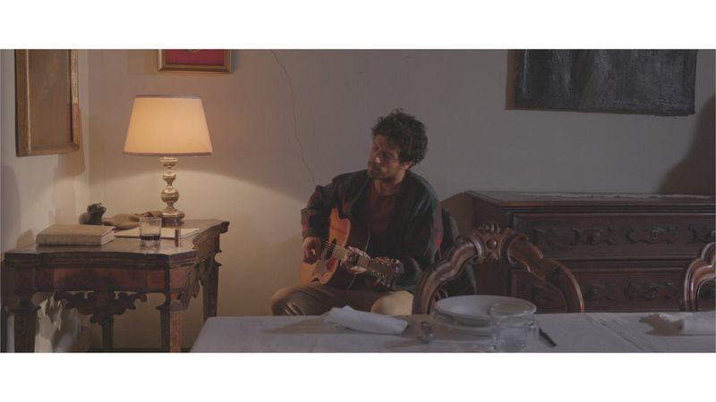 A man playing a guitar, sitting in a dining room with ornate but ageing wooden furniture.