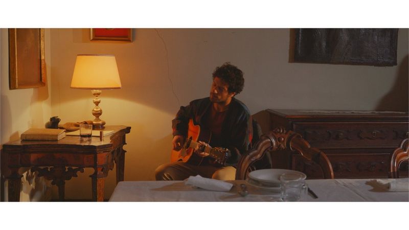 A man playing a guitar, sitting in a dining room with ornate but ageing wooden furniture. The image has been colour graded in post-production.