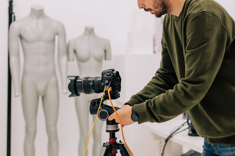 A man adjusts a Canon EOS camera on a tripod in a photography studio, with an external power supply connected and a tethering cable running from its USB port. 