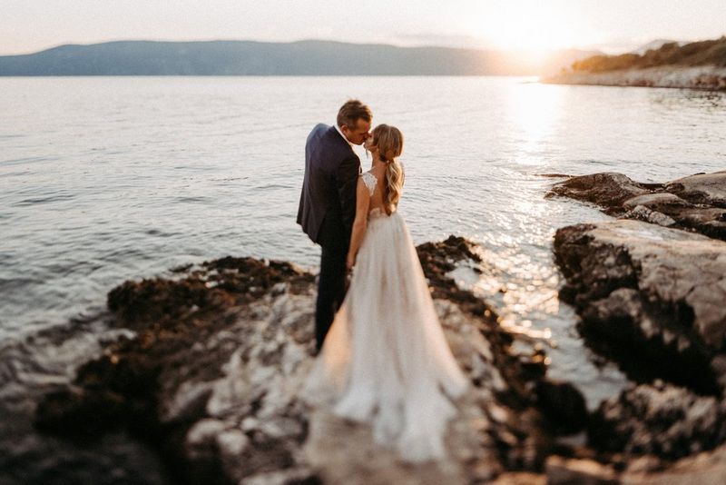 A bride and groom in their wedding attire kiss as they stand on rocks with a body of water and mountains visible in the background. Shot on a Canon EOS R5 by Carmen and Ingo Photography.
