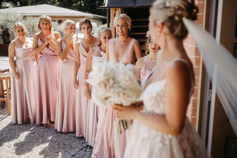 Seven bridesmaids look at a bride holding a bouquet of flowers, shot on a Canon EOS R6 by Carmen and Ingo Photography.  