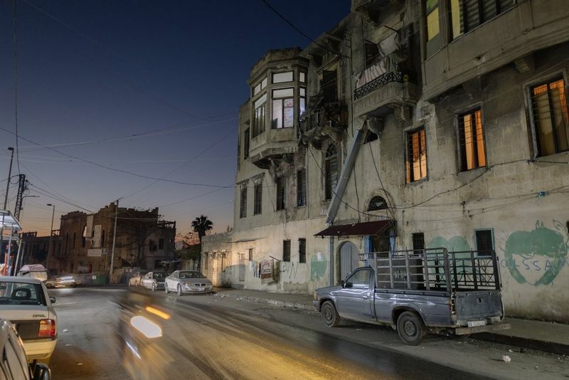 A street scene is captured at night, with buildings illuminated from within and cars parked either side of the road, in a photo by Aline Deschamps taken on a Canon EOS R8.