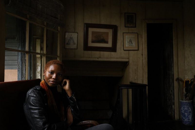 A woman in a leather jacket sits in a darkened room, her face partially lit by natural light from the window behind her.