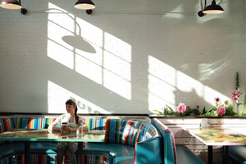 A woman seated at a table on a padded, rainbow-striped bench, with the shadow of the window frame filling the wall behind her.