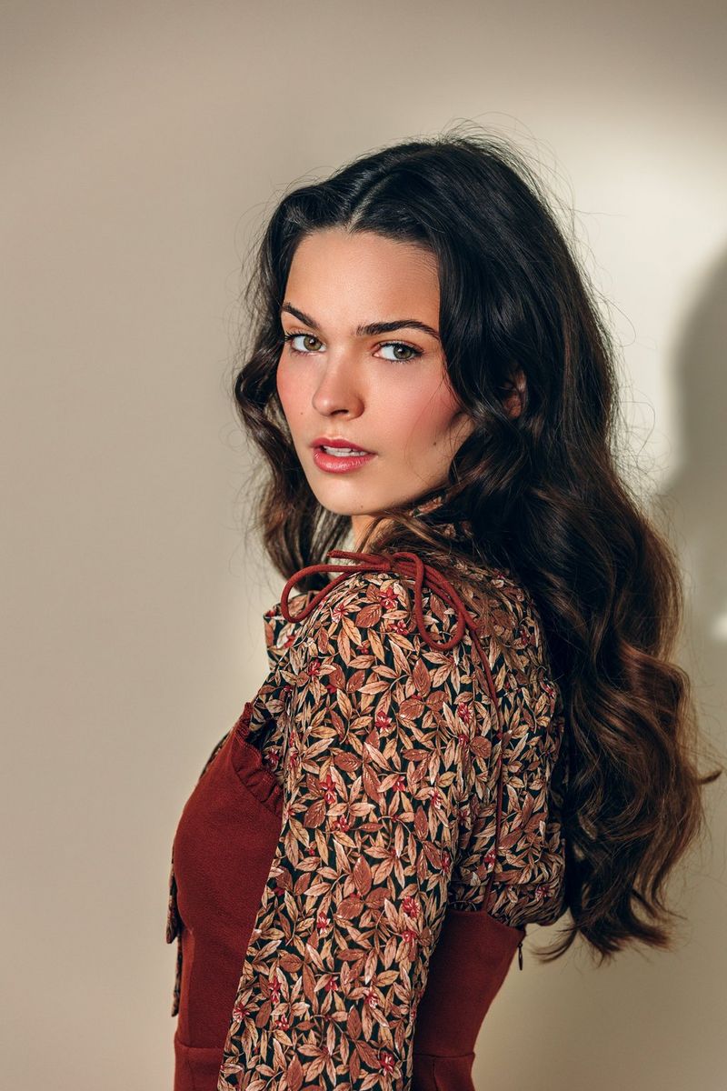 A model wearing a dress and printed blouse stands side on to the camera, turning her head to look at it.
