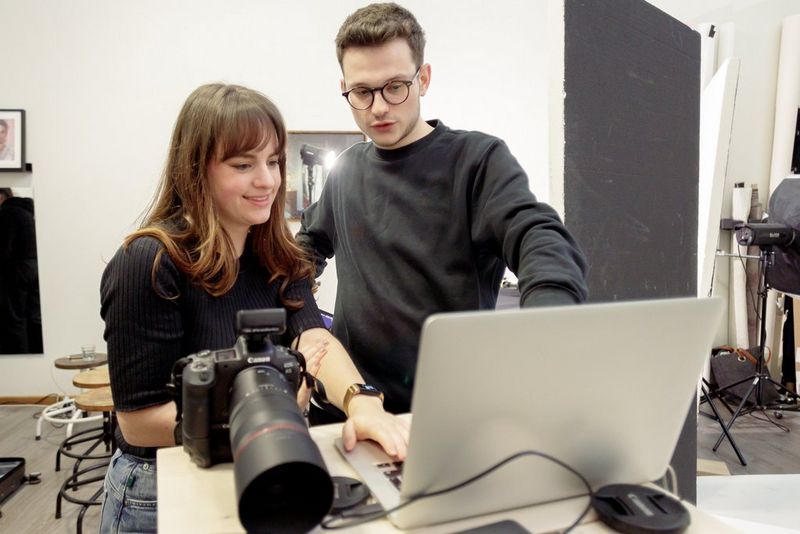 Two people stand in front of a laptop, both looking at the screen, with a Canon EOS R5 on the table next to it.
