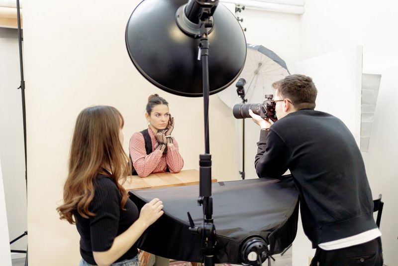 A female model poses at a high tabletop underneath a beauty dish. A male photographer leans in to take a shot, and a woman stands to the side offering direction.