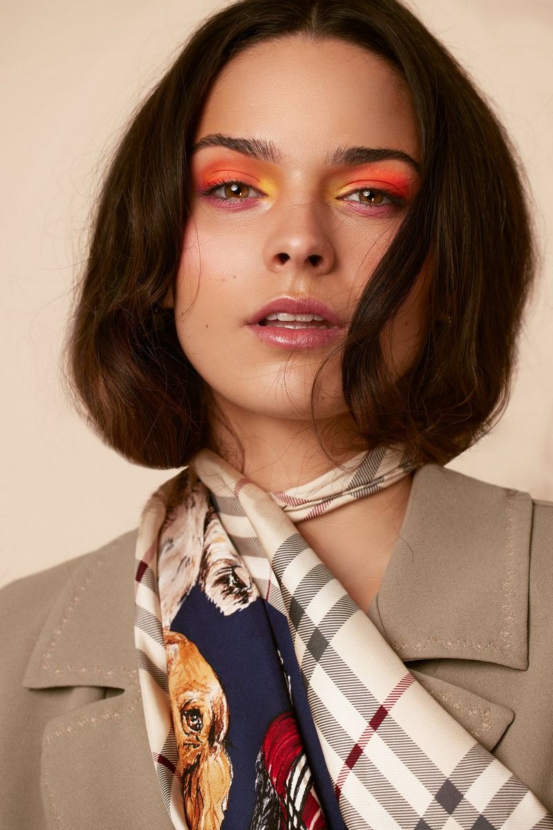 A close-up of a model in colourful eye make-up, a jacket and a printed scarf around her neck, looking at the camera.