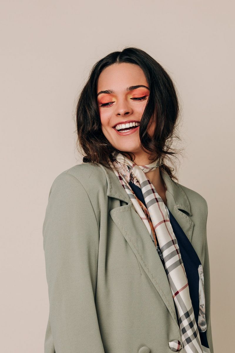 A model in colourful eye make-up, a jacket and a printed scarf around her neck, laughing and looking down to the side.