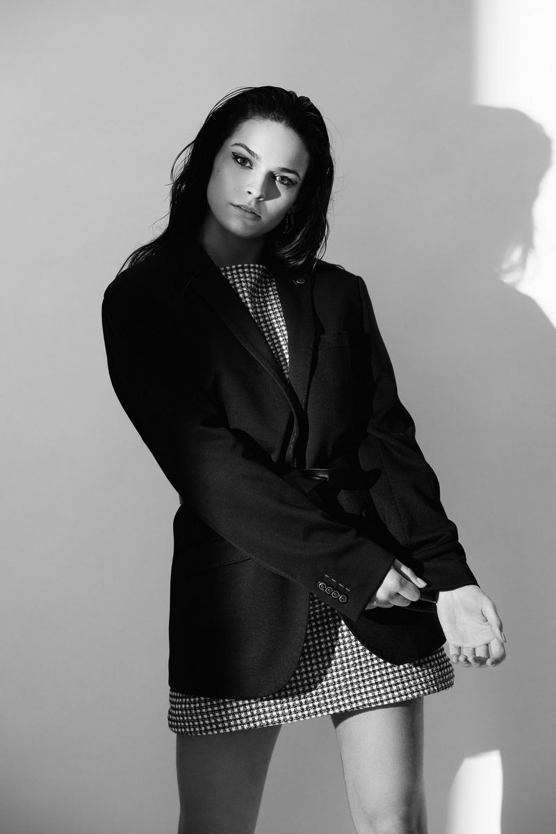 A fashion model, shot from the knees upwards, wearing a long black jacket over a short printed dress, and lit so her profile is just visible as a shadow on the pale background.