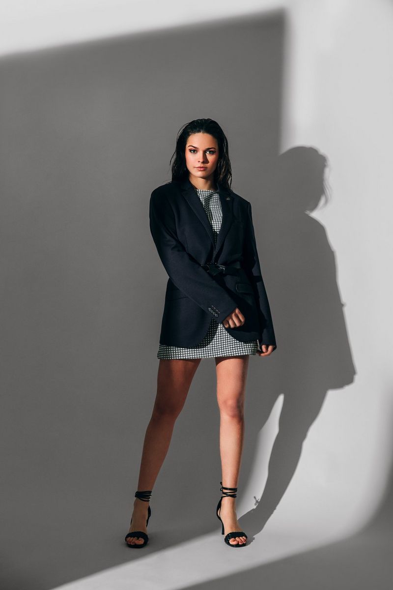 A full-length shot of a fashion model wearing a long black jacket over a short printed dress, lit so her pose casts a shadow on the pale background.