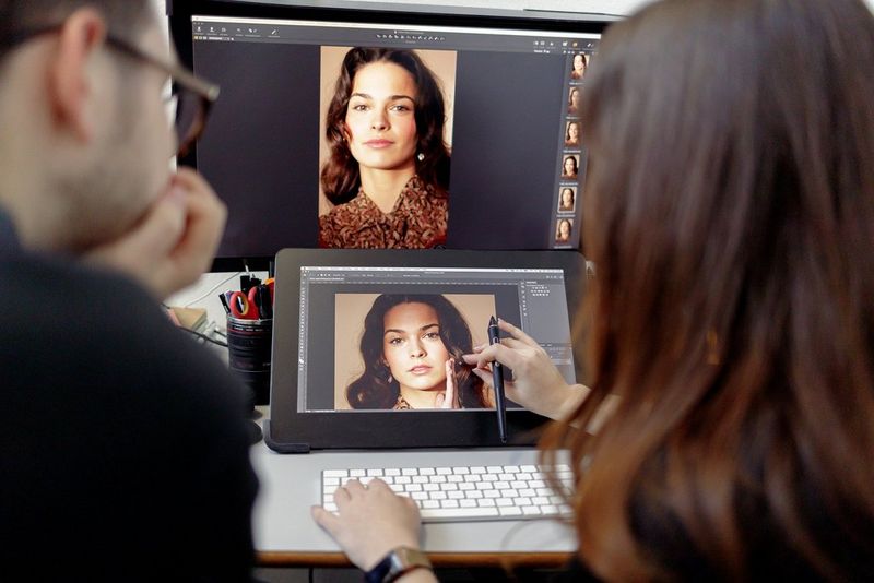 A person edits an image of a model on their laptop, while another person watches them. Behind the laptop is a large monitor showing a different image of the same model.
