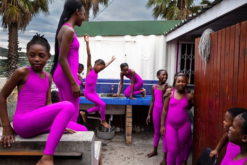 A group of young girls in identical pink leotards and leggings mingle outside a small building. Taken by Brent Stirton on a Canon EOS R5.